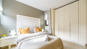 Un lit bien fait, avec une literie blanche et un seul oreiller jaune, une table de chevet avec une lampe et une armoire aux portes coulissantes.