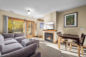 Una sala de estar con un sofá gris, una chimenea, una mesa de comedor con sillas y un televisor sobre la chimenea.
