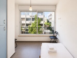 A hotel room with a large window, a bed with white linens, a black chair, and a hanging light fixture. - Bright 2BR in Hype Neighborhood by FeelHome (Tel Aviv - Jaffa)