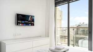 Une chambre d’hôtel moderne, dotée d’un grand téléviseur à écran plat fixé au mur et diffusant un film. On y trouve un lit avec du linge de lit blanc et deux serviettes pliées posées sur le rebord de la fenêtre. La chambre dispose d’une grande fenêtre offrant une vue sur le paysage urbain.