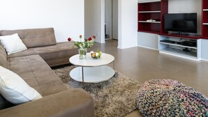 A living room with a brown sofa, a round coffee table with a vase of flowers, and a flat-screen TV on a red and white entertainment unit. - MAMAD Comfort 3BR & Balcony in City Center by FeelHome (Tel Aviv - Jaffa)