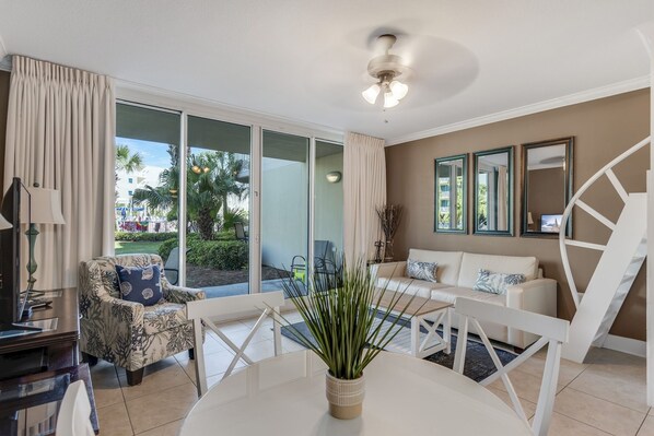 A modern living room with a glass door leading to a balcony, a white sofa, a patterned armchair, and a dining table with a plant centerpiece.