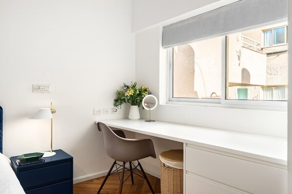 Un dormitorio con un escritorio blanco, una silla gris, una mesita de noche con una lámpara, una planta en la mesita de noche y una ventana con vistas a edificios.