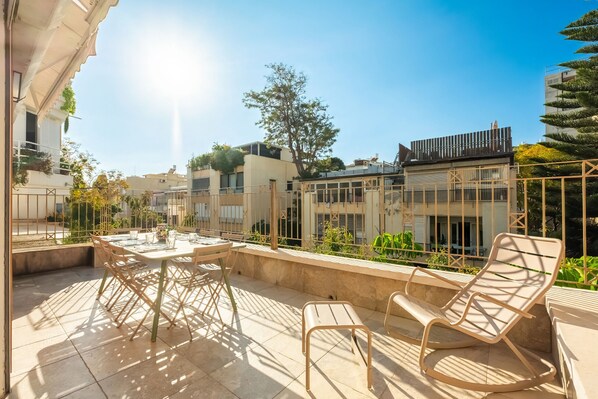 A terrace with a table and chairs, surrounded by a low fence and greenery.
