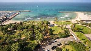 Vista aérea de una zona costera con un puerto deportivo, playa y parque con árboles y senderos.