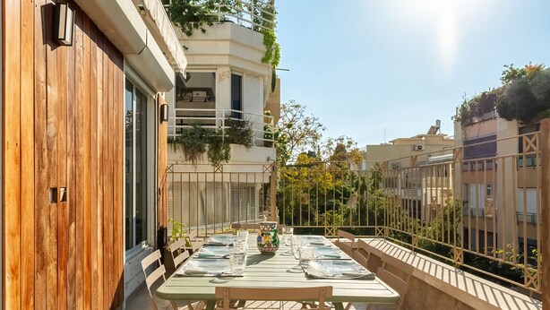 Un balcón con una mesa lista para dos, una puerta de madera, y un edificio con balcones y follaje.