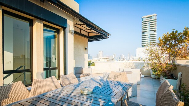 A rooftop patio with a wooden table and wicker chairs, overlooking a city skyline.