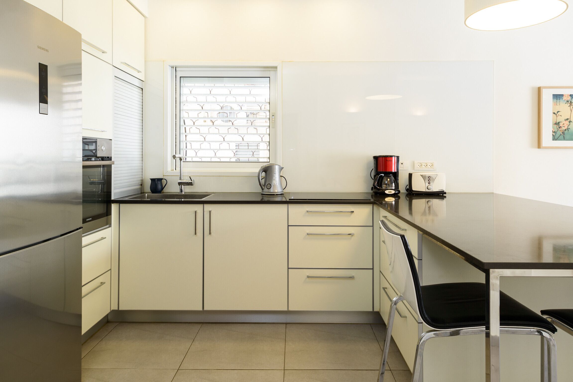A modern kitchen with a stainless steel refrigerator, a kettle, a coffee maker, and a window with blinds.