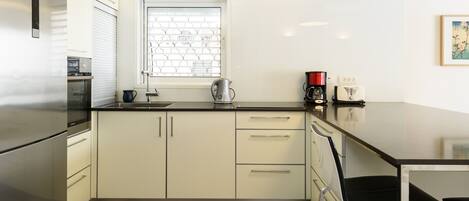 A modern kitchen with a stainless steel refrigerator, a kettle, a coffee maker, and a window with blinds.