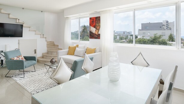 A modern living room with a staircase, a flat-screen TV, a dining table with chairs, and large windows offering a city view. - Penthouse with Terrace & Panoramic View by FeelHome (Tel Aviv - Jaffa)