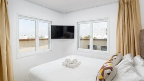 A hotel room with a bed, two windows, a flat-screen TV, and a city view. - Penthouse with Terrace & Panoramic View by FeelHome (Tel Aviv - Jaffa)
