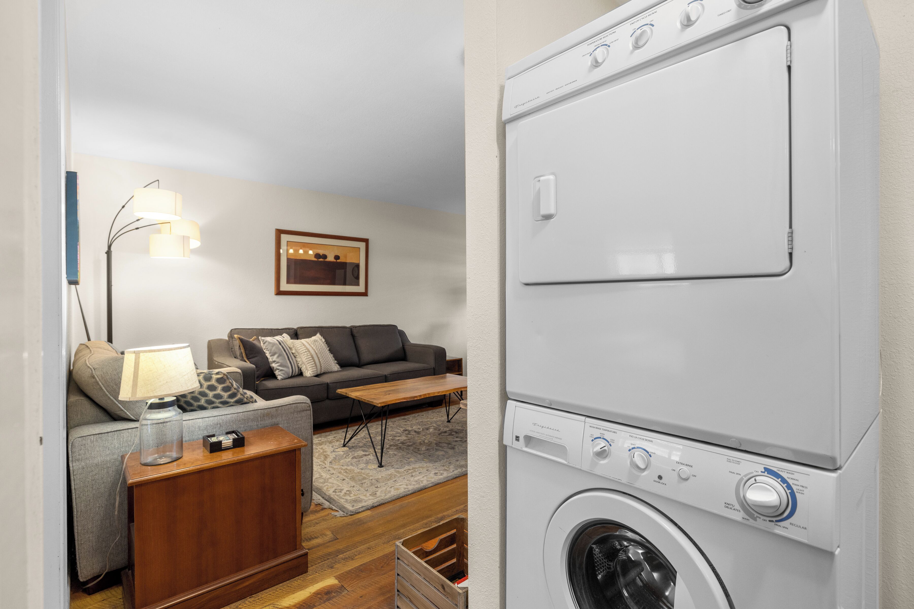Une buanderie équipée d’un lave-linge et d’un sèche-linge à chargement frontal, d’une armoire en bois, d’une lampe et d’un canapé gris agrémenté d’un coussin décoratif à motifs.