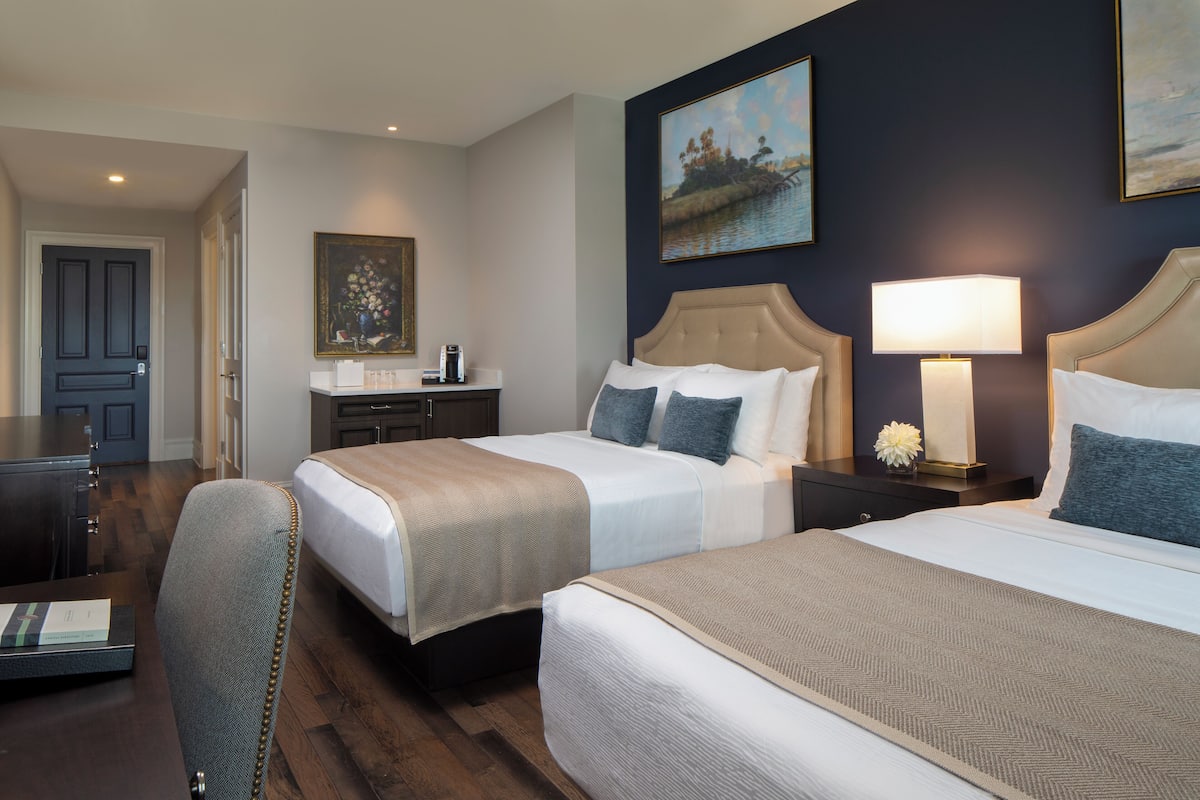 A hotel room with two beds, a desk, and a chair. There are paintings on the walls and a lamp on the nightstand.