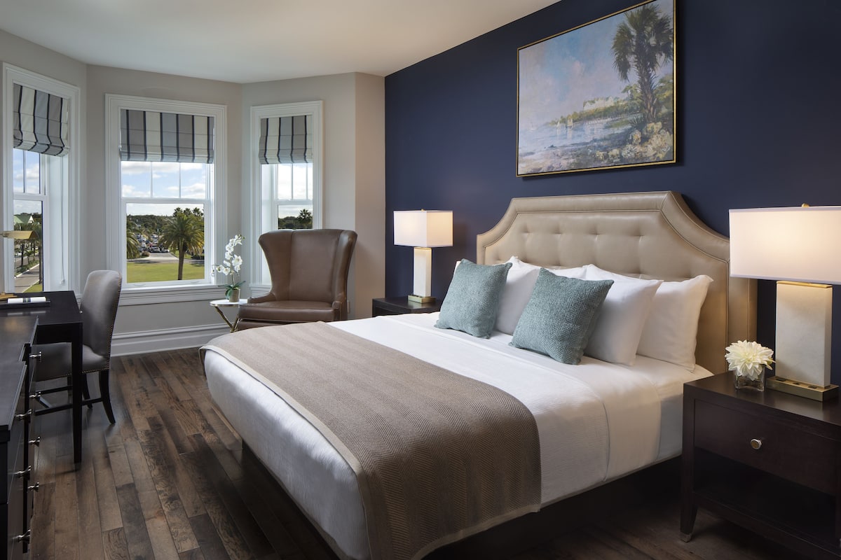A bedroom with a large bed, two bedside lamps, a chair, a desk, and a painting on the wall.