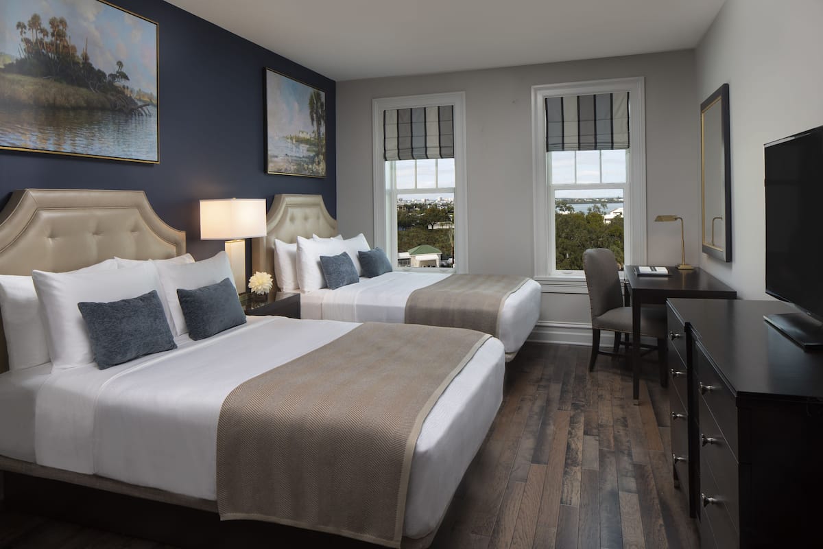 A hotel room with two beds, a desk, a chair, and a TV.
