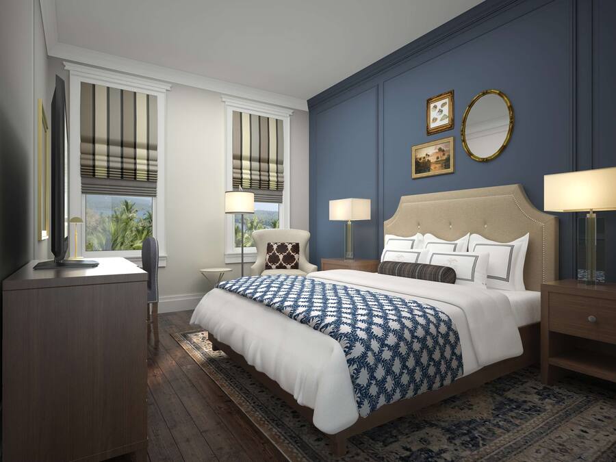 A bedroom with a large bed, two bedside tables, a nightstand, a desk, and a chair. There are two windows with blinds, a wall-mounted mirror, and two framed pictures.