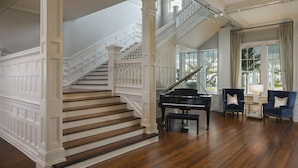 A grand staircase with white balustrades leading to a second floor, a grand piano, and two armchairs in a spacious room with hardwood flooring. - Belleview Inn (Belleair)