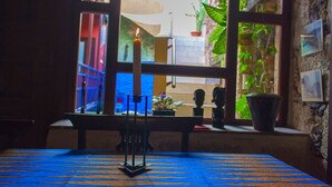 A room with a table covered in a patterned tablecloth, a window with a view of a street and greenery, and a wall with framed pictures. - Casa Luna (Agaete)