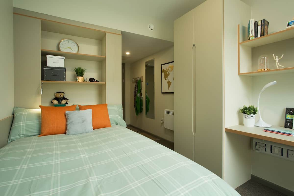 A bedroom with a bed, pillows, a clock, a shelf with books, a globe, and a desk with a lamp.