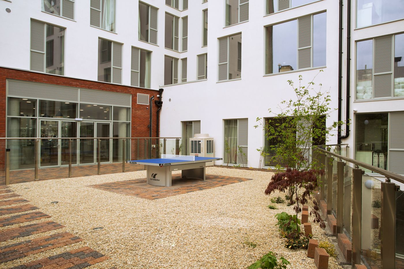 A courtyard with a ping pong table, a brick wall, and a modern building with large windows and a glass railing.