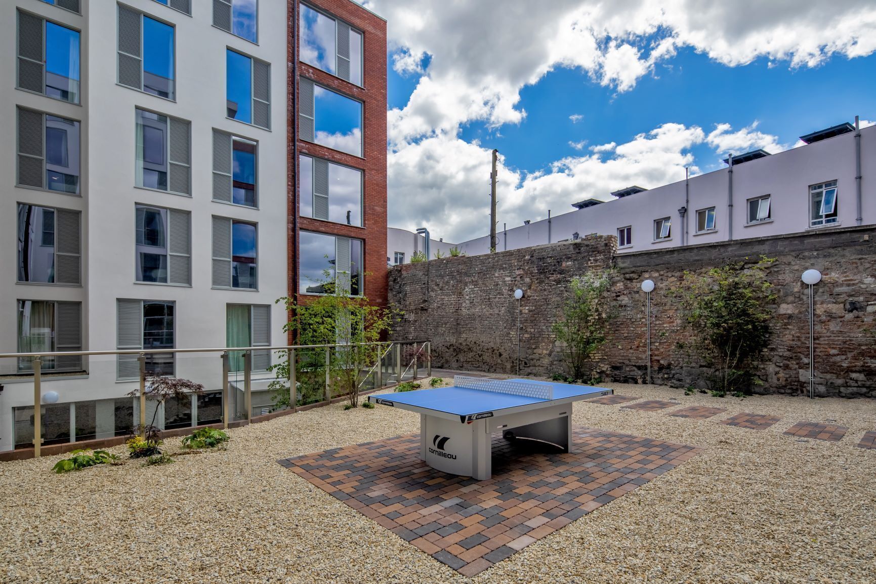 A ping pong table in an outdoor courtyard with a gravel and brick floor, surrounded by buildings with large windows and a stone wall.