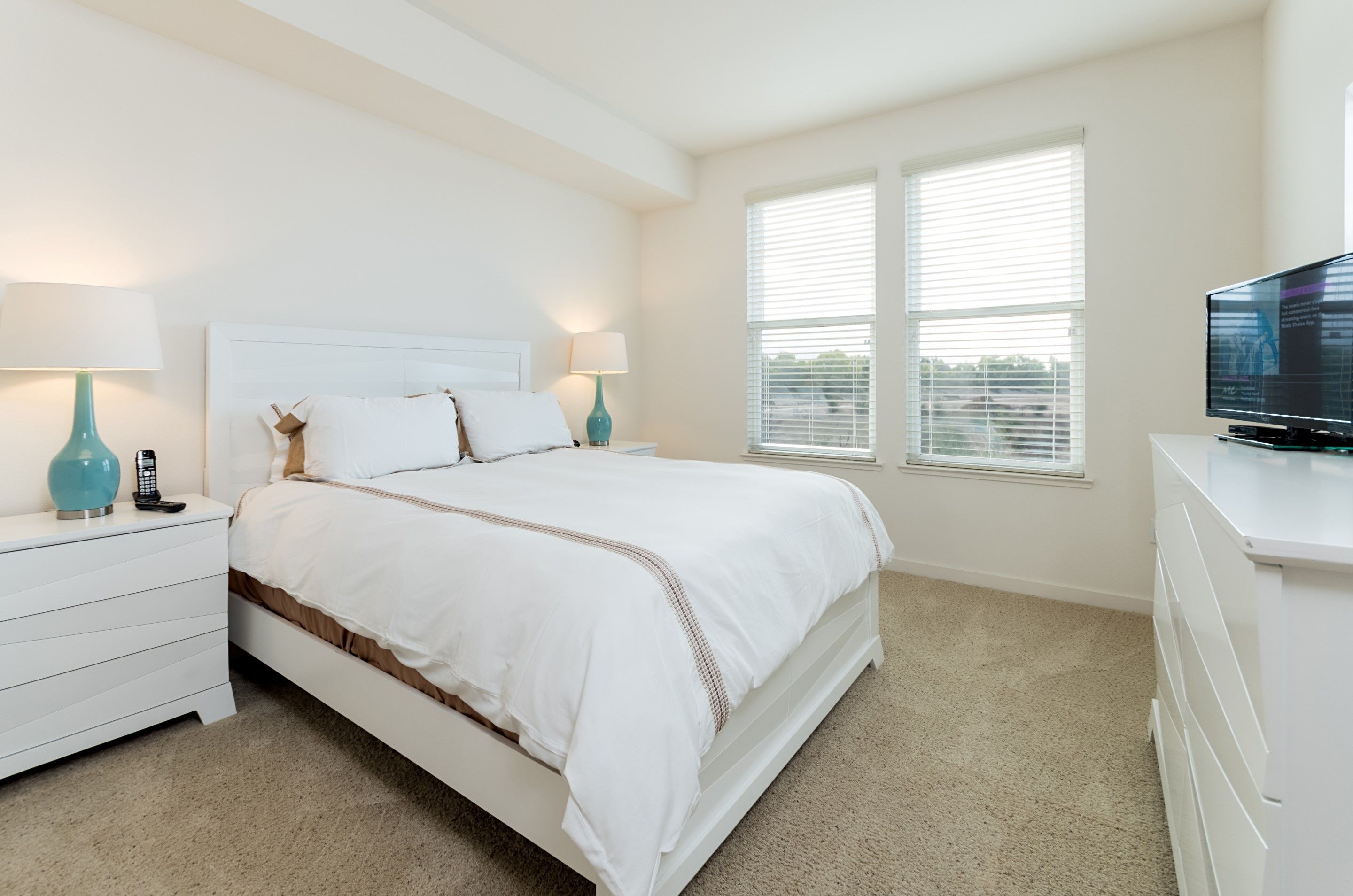 A bedroom with a white bed, two bedside tables with lamps, a TV on a dresser, and a window with blinds.