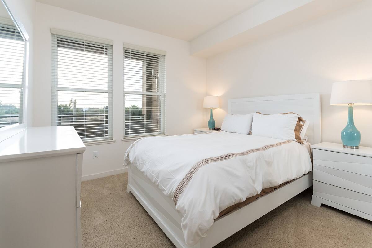 A bedroom with a bed, white nightstands, teal lamps, and a window with blinds.