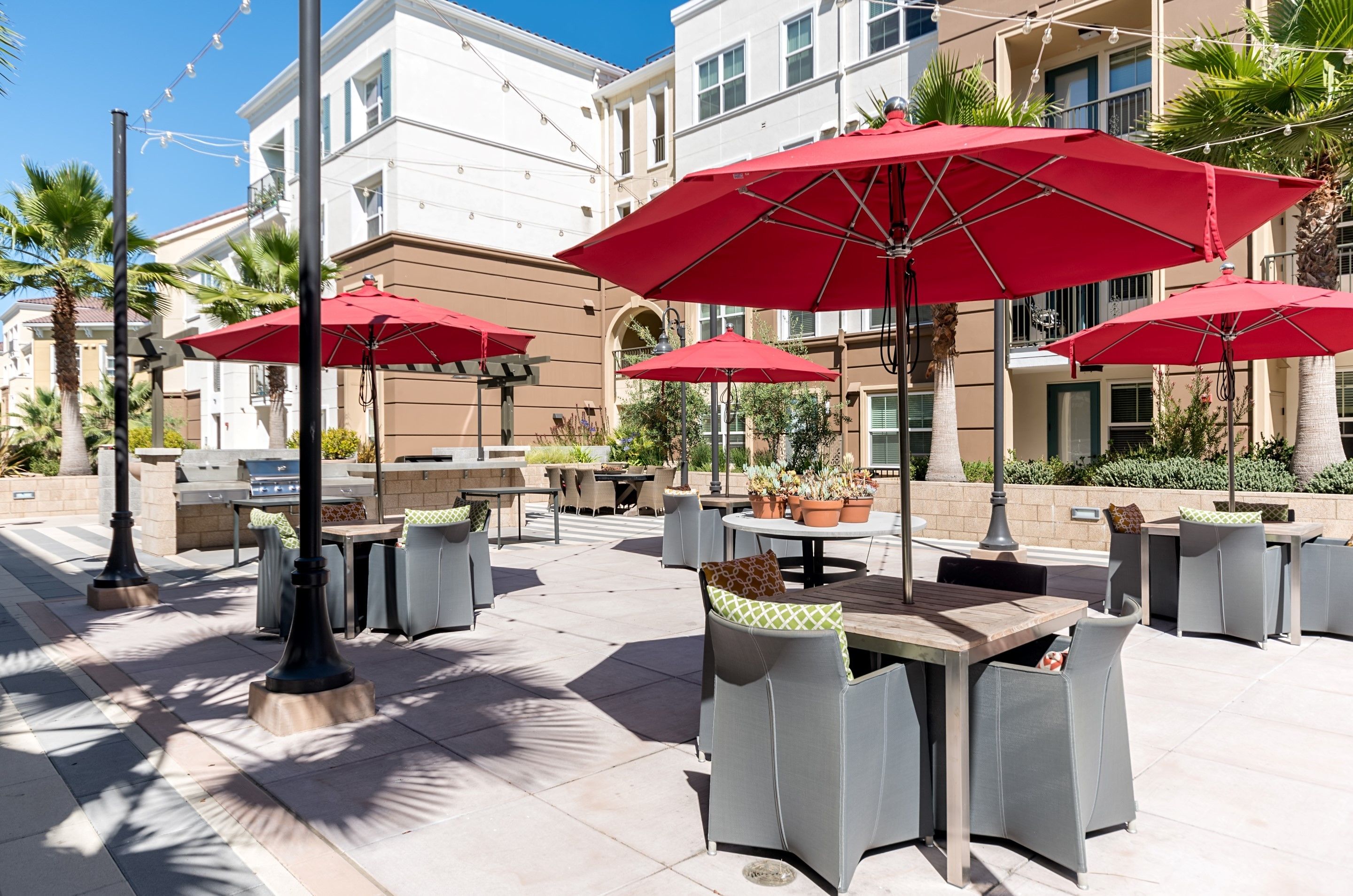 An outdoor patio area with tables and chairs, red umbrellas, and potted plants.
