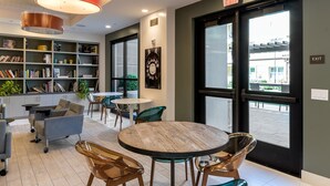 A modern indoor space with a round wooden table, chairs, a bookshelf, and a large window. - Bluebird Suites in Silicon Valley (San Jose)
