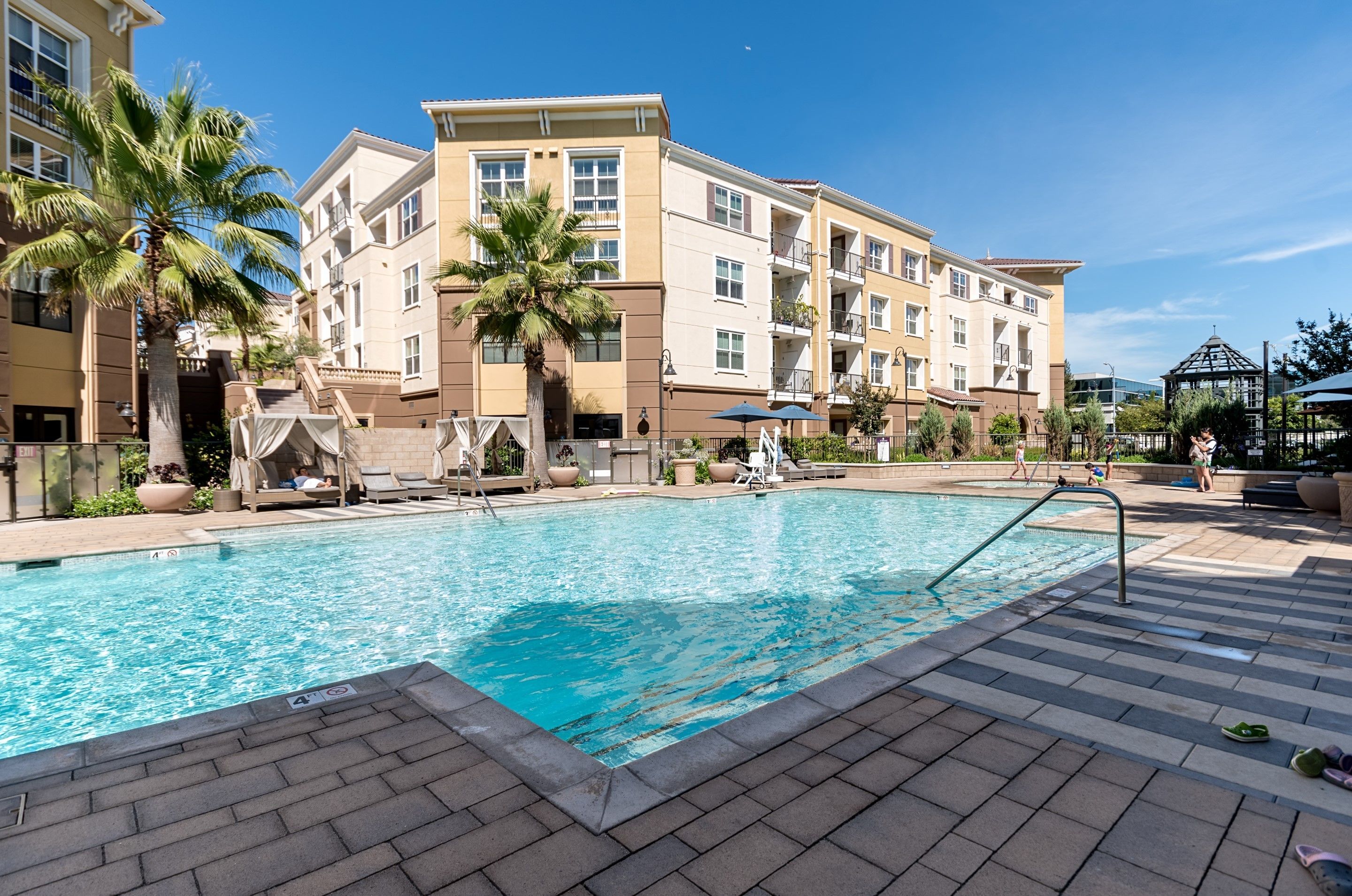 A swimming pool with lounge chairs and palm trees, surrounded by multi-story buildings with balconies.