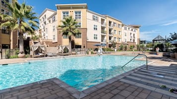 A swimming pool with lounge chairs and palm trees, surrounded by multi-story buildings with balconies.