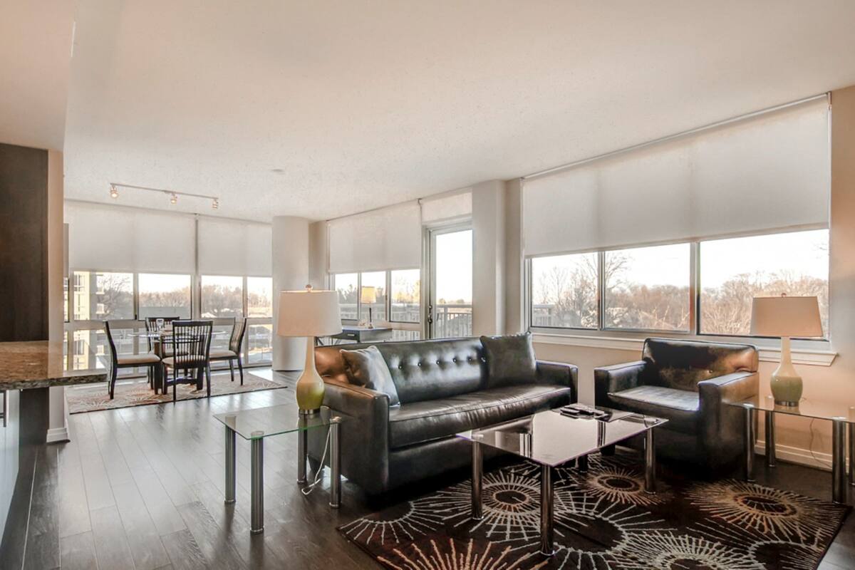 A spacious living room with a black leather sofa, a glass coffee table, and a patterned rug. There is a dining area with a table and chairs, and large windows providing a view of the outdoors.