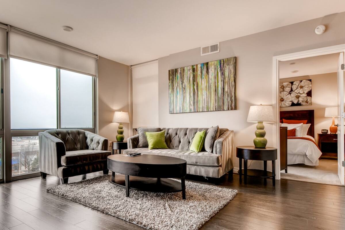 A modern living room with a sofa, armchair, and coffee table, a large window, and a painting on the wall.