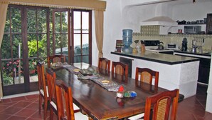A dining area with a wooden table and chairs, a kitchen with a water dispenser, and a view of greenery through large windows. - Casita Jazmin (Antigua Guatemala)