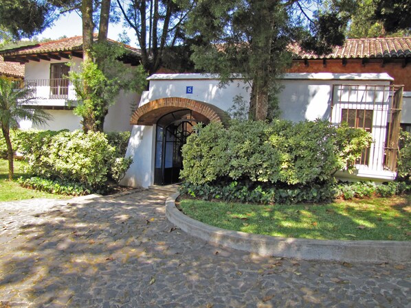 A building with a tiled roof, a stone pathway leading to an arched entrance, and a well-maintained garden with various shrubs. - Casita Jazmin (Antigua Guatemala)