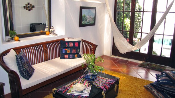 A living room with a sofa, a hammock, a table with a plant, and a window with a view of greenery. - Casita Jazmin (Antigua Guatemala)