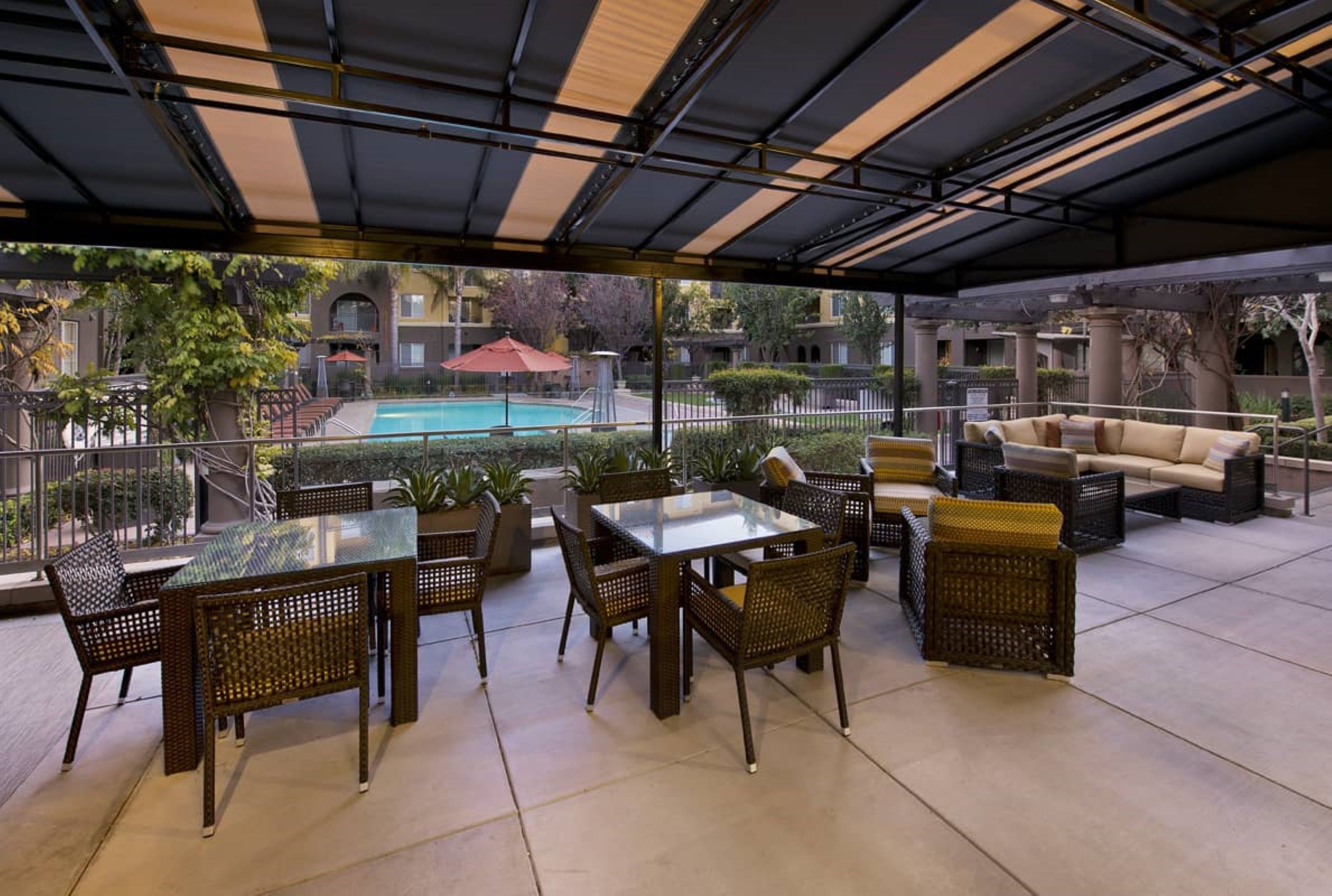 An outdoor patio area with a glass table and wicker chairs, overlooking a swimming pool and a building with balconies.