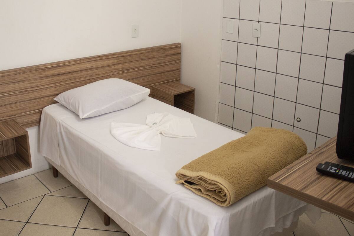 A single bed with a wooden headboard, a white pillow, a folded white towel, and a brown towel. A remote control is on a wooden nightstand beside the bed.