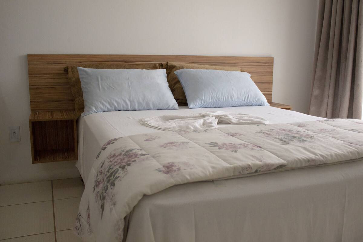 A neatly made bed with a floral bedspread and two pillows, a wooden headboard, and a wooden nightstand with a lamp.