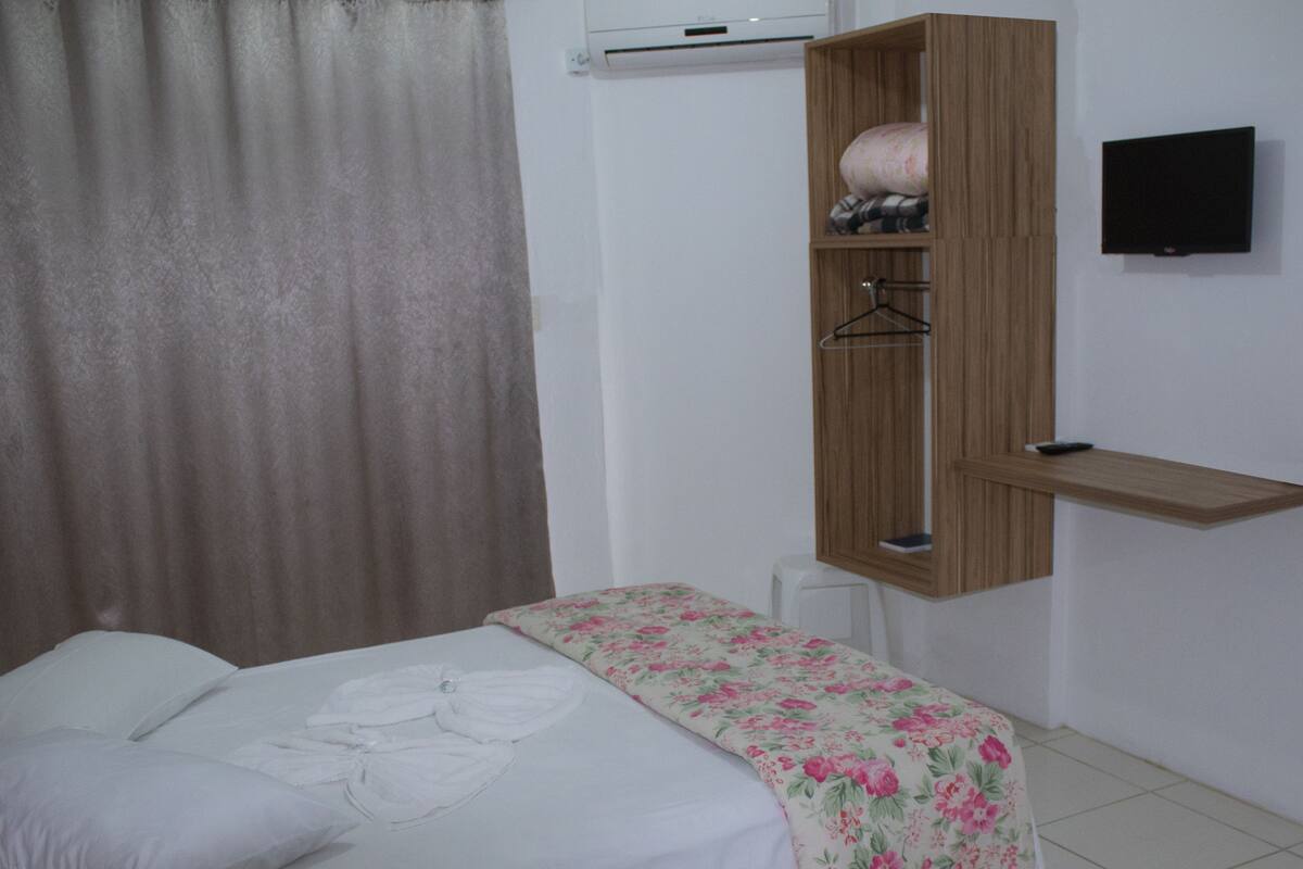 A neatly made bed with a floral bedspread, a wooden wardrobe with a hanging shelf, and a mounted television on the wall.