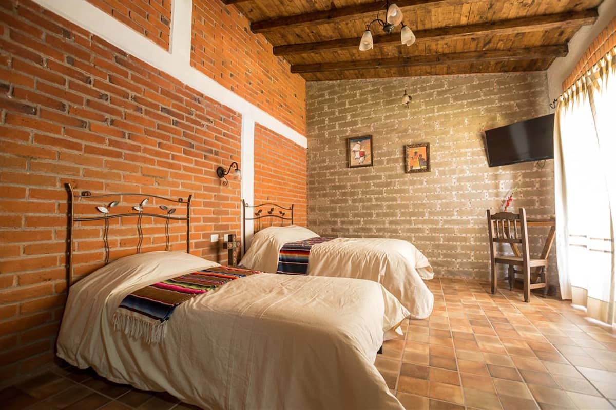 A room with two beds, a brick wall, a wooden ceiling, and a TV mounted on the wall.