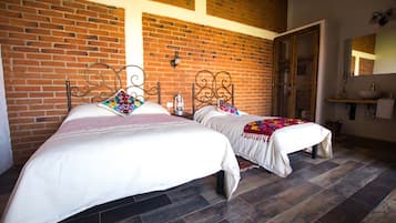 A room with two beds, a brick wall, a wooden floor, and a bathroom with a sink and mirror.
