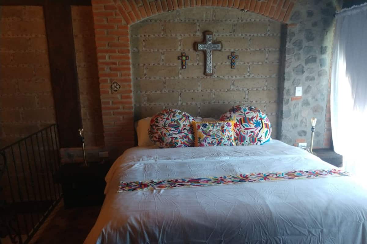 A bedroom with a brick wall, a large bed with colorful pillows, and a cross on the wall.