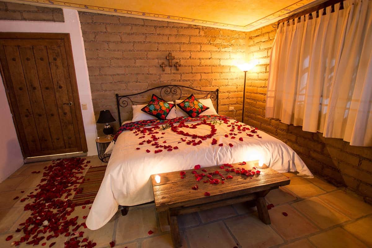 A cozy bedroom with a brick wall, a wooden door, a bed with rose petals, and a bench.