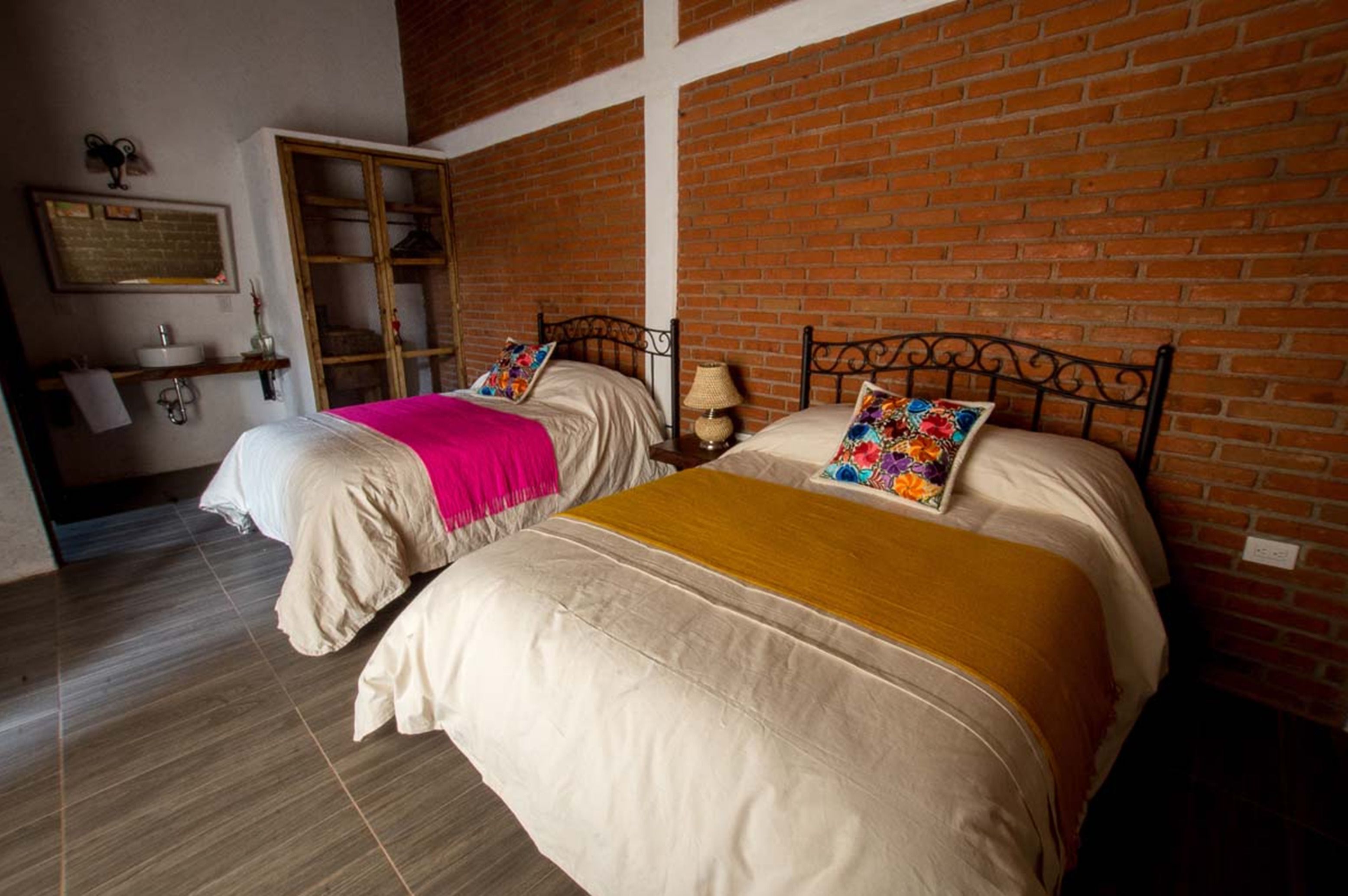 Quarto duplo com duas camas, cada uma com cabeceira de madeira, parede de tijolos e pequena mesa de cabeceira com luminária.