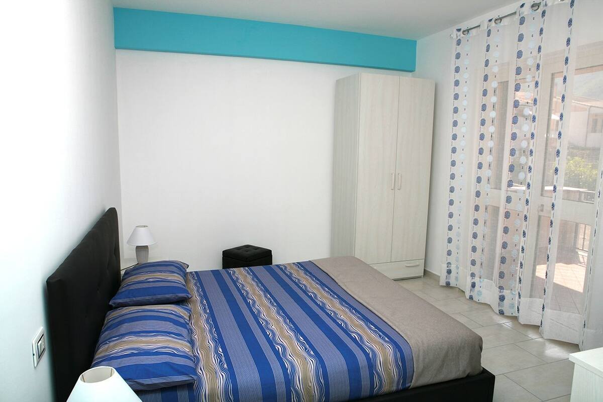 A bedroom with a bed, a nightstand, a lamp, a wardrobe, and a view through a window with curtains.