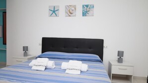 Una cama bien hecha con ropa de cama azul y blanca a rayas, dos toallas blancas y un cabecero tapizado negro. Hay tres obras de arte en la pared que representan estrellas de mar y conchas marinas.