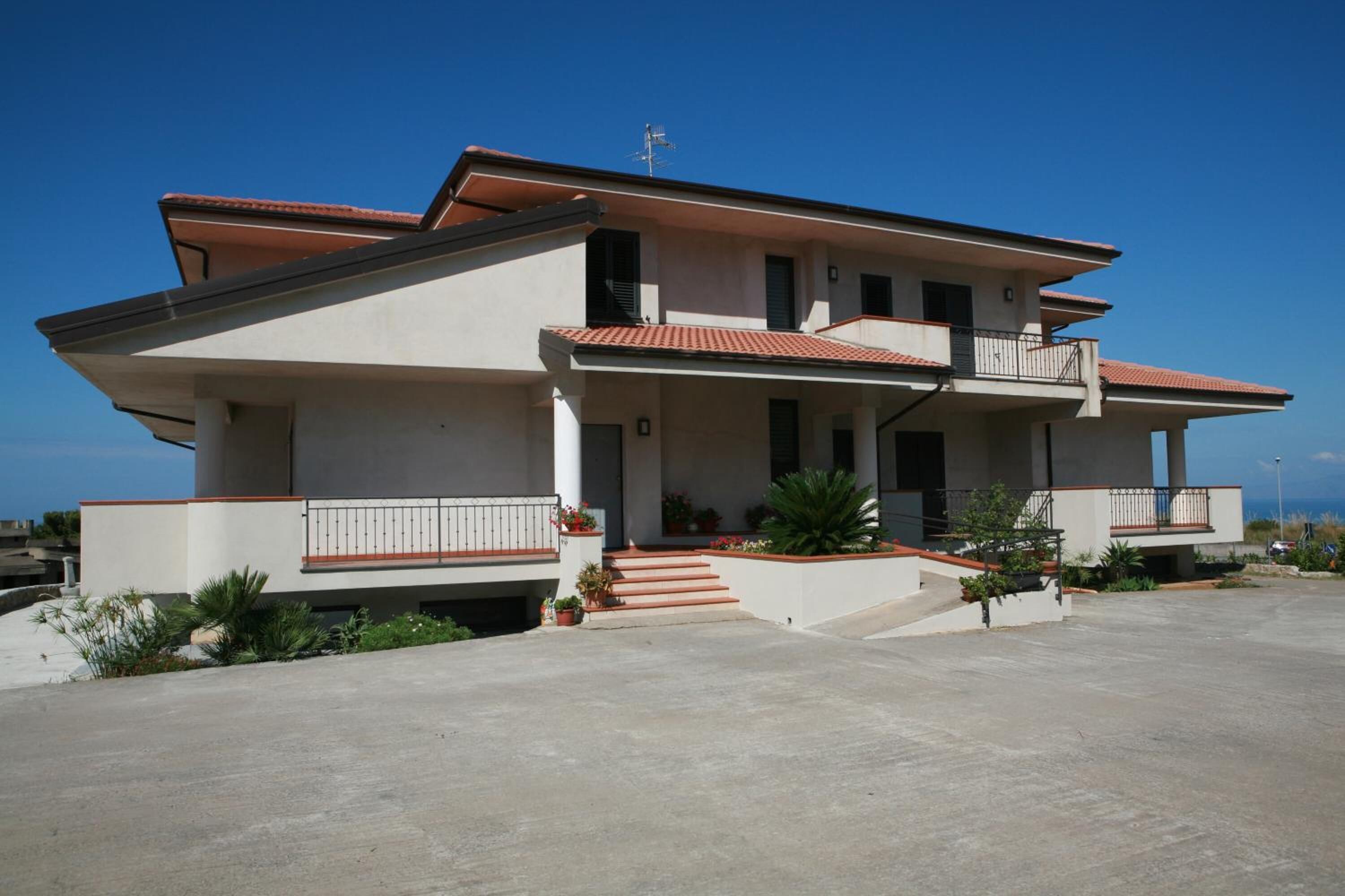 A two-story house with a balcony, a red-tiled roof, and a small garden area.