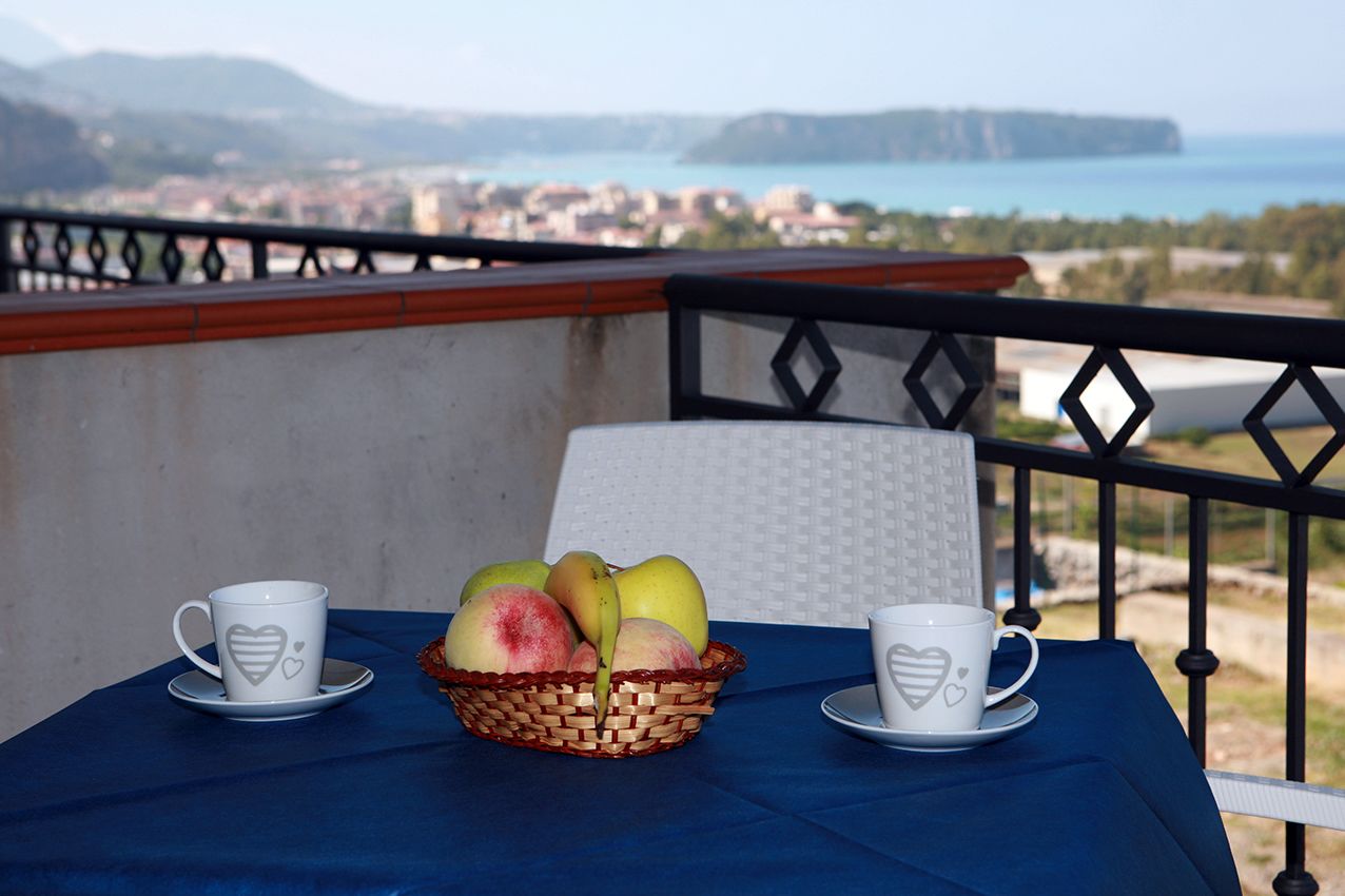 A balcony with a table set for two, featuring a basket of fruit and two heart-shaped cups.