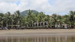 A beachfront hotel with multiple-story buildings, palm trees, and a mountain in the background. - Club del cielo Unit 1 (Jacó)